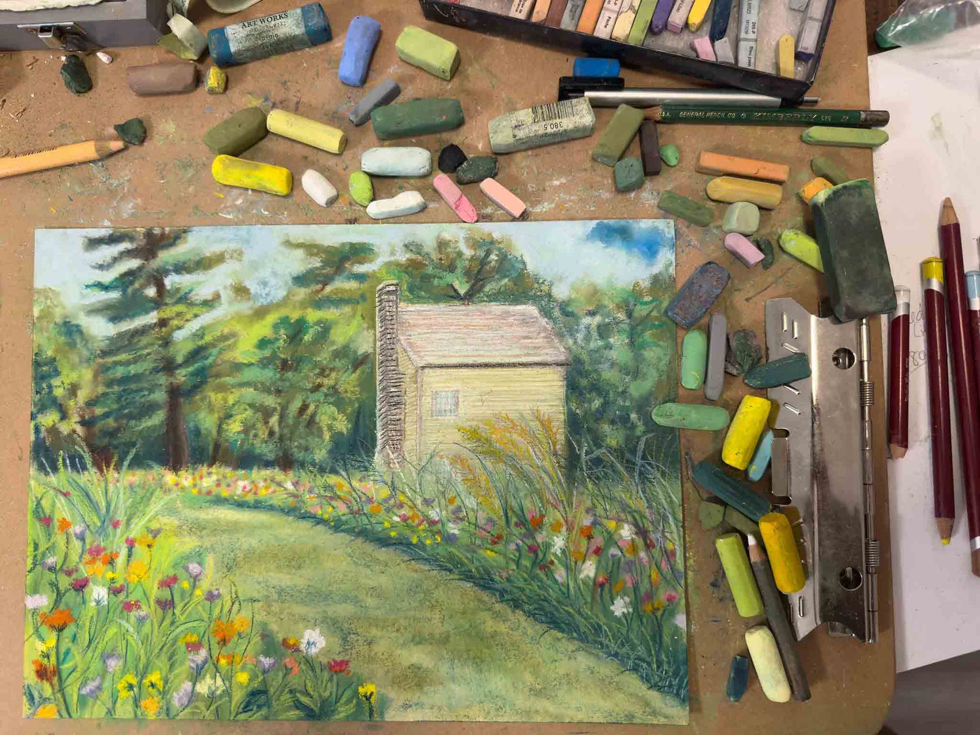 Soft pastel pieces on an art board surrounding a pastel painting of a Shaker garden and Shaker building.