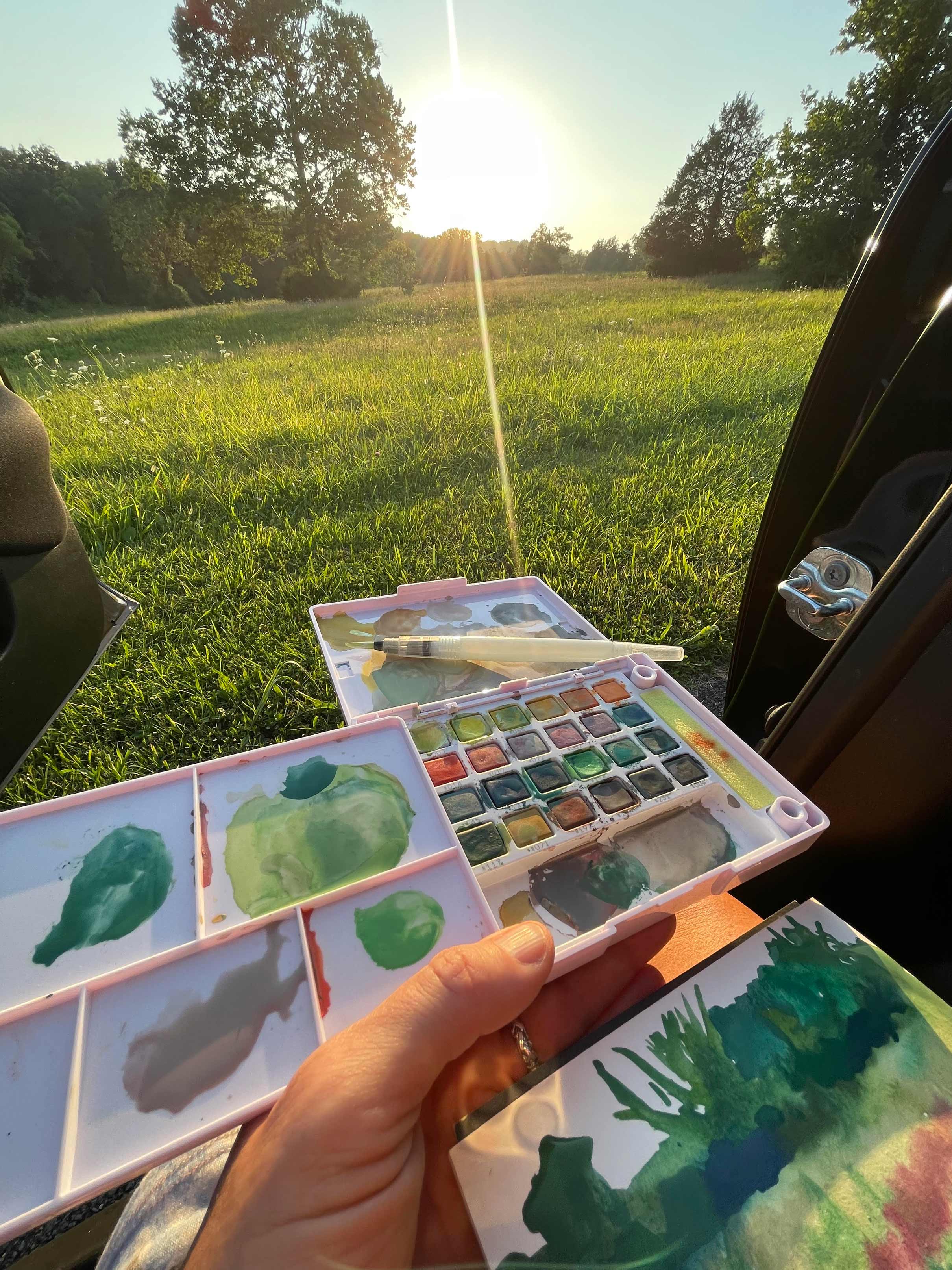 A photo of the artist painting from her car, holding a travel set of watercolors while working on the painting.